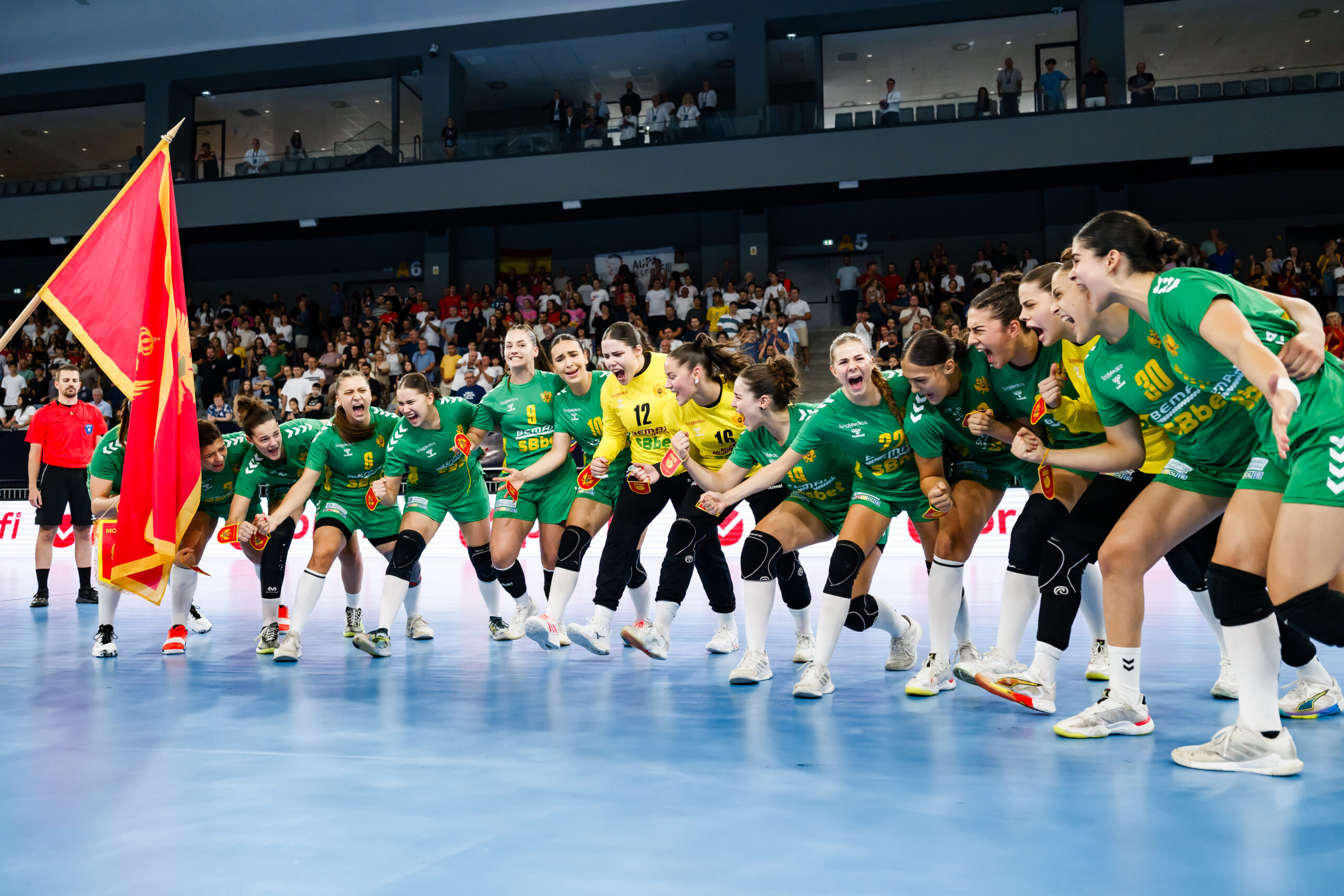 A team in green uniforms celebrates on a blue court with a red flag, surrounded by cheering spectators.